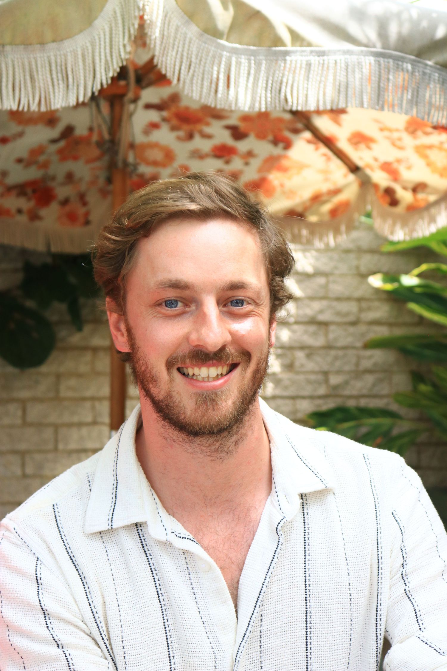 Smiling young bearded man in striped white shirt posing outdoors under decorative fringed umbrella.