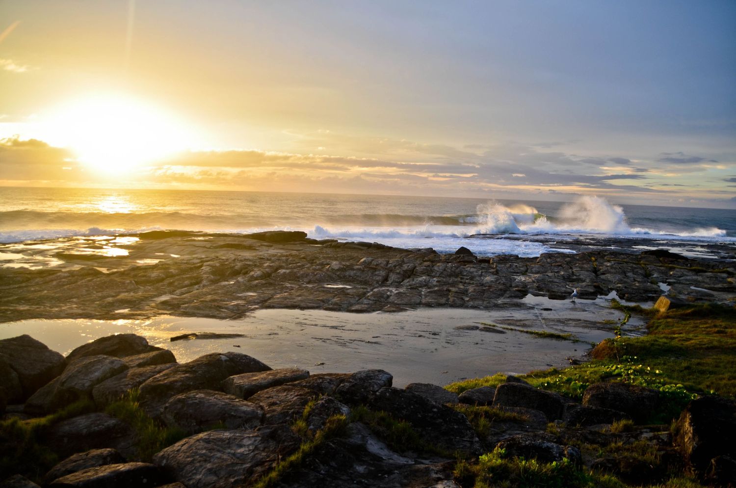 Dramatic sunset over rocky coastline with crashing waves and golden sky.