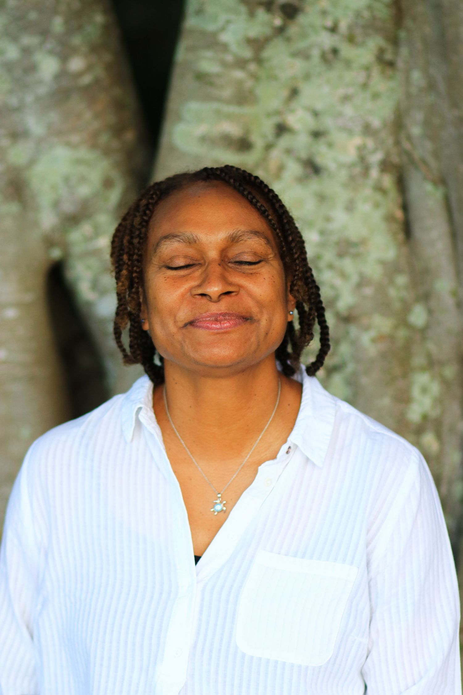 Peaceful middle-aged Black woman with braids wearing white shirt meditating with eyes closed outdoors.