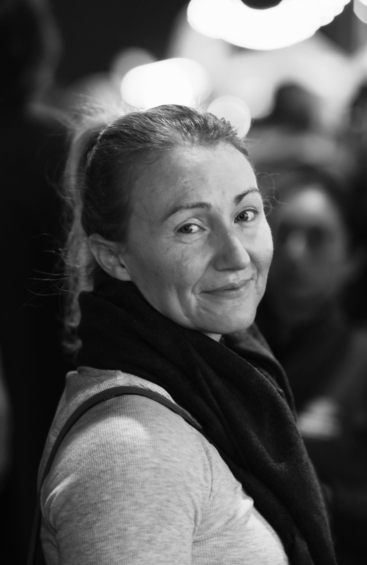 Black and white close-up portrait of a smiling woman with a scarf in a busy setting.