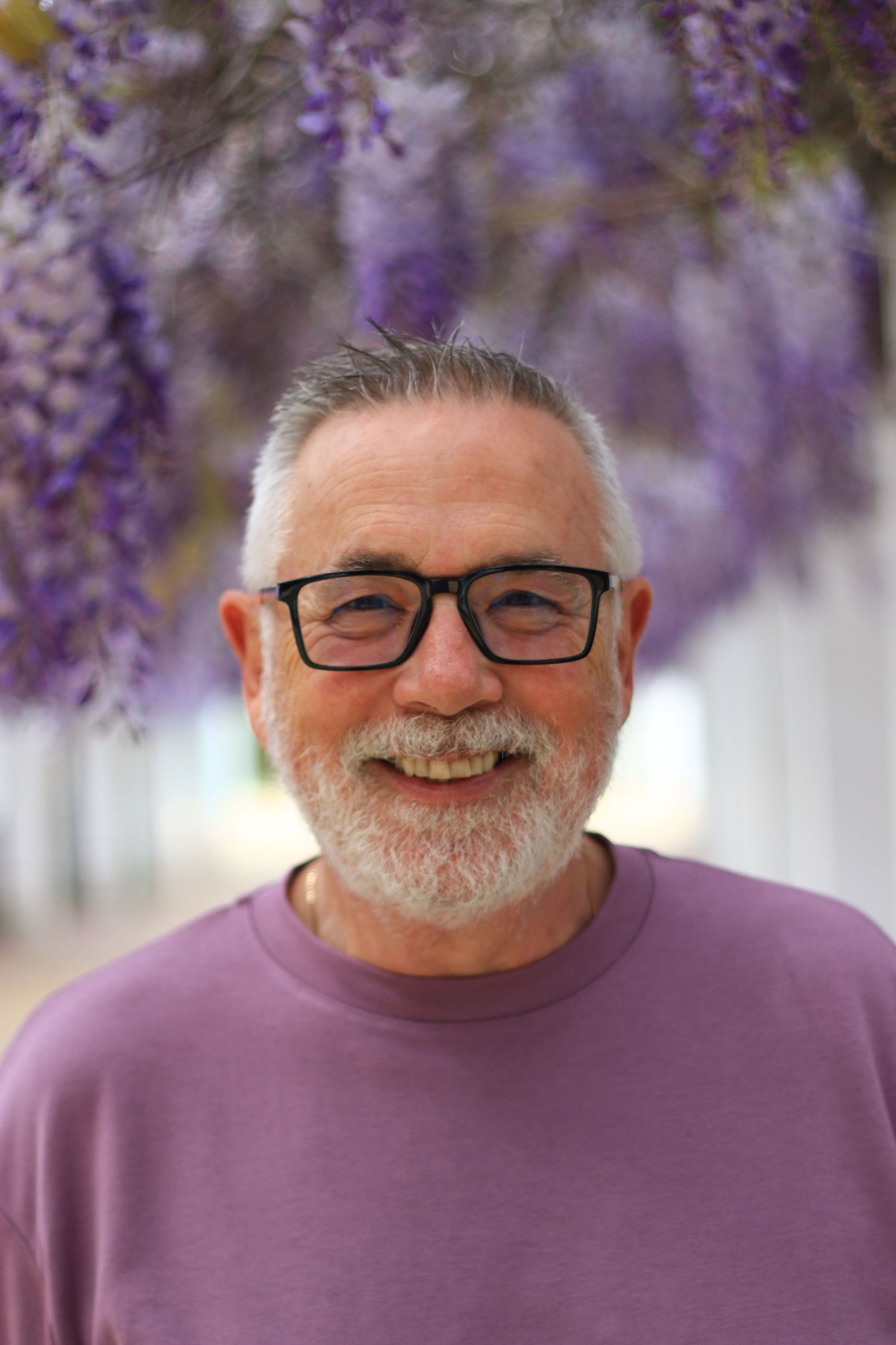 Smiling older man with gray beard and glasses standing under purple wisteria flowers.