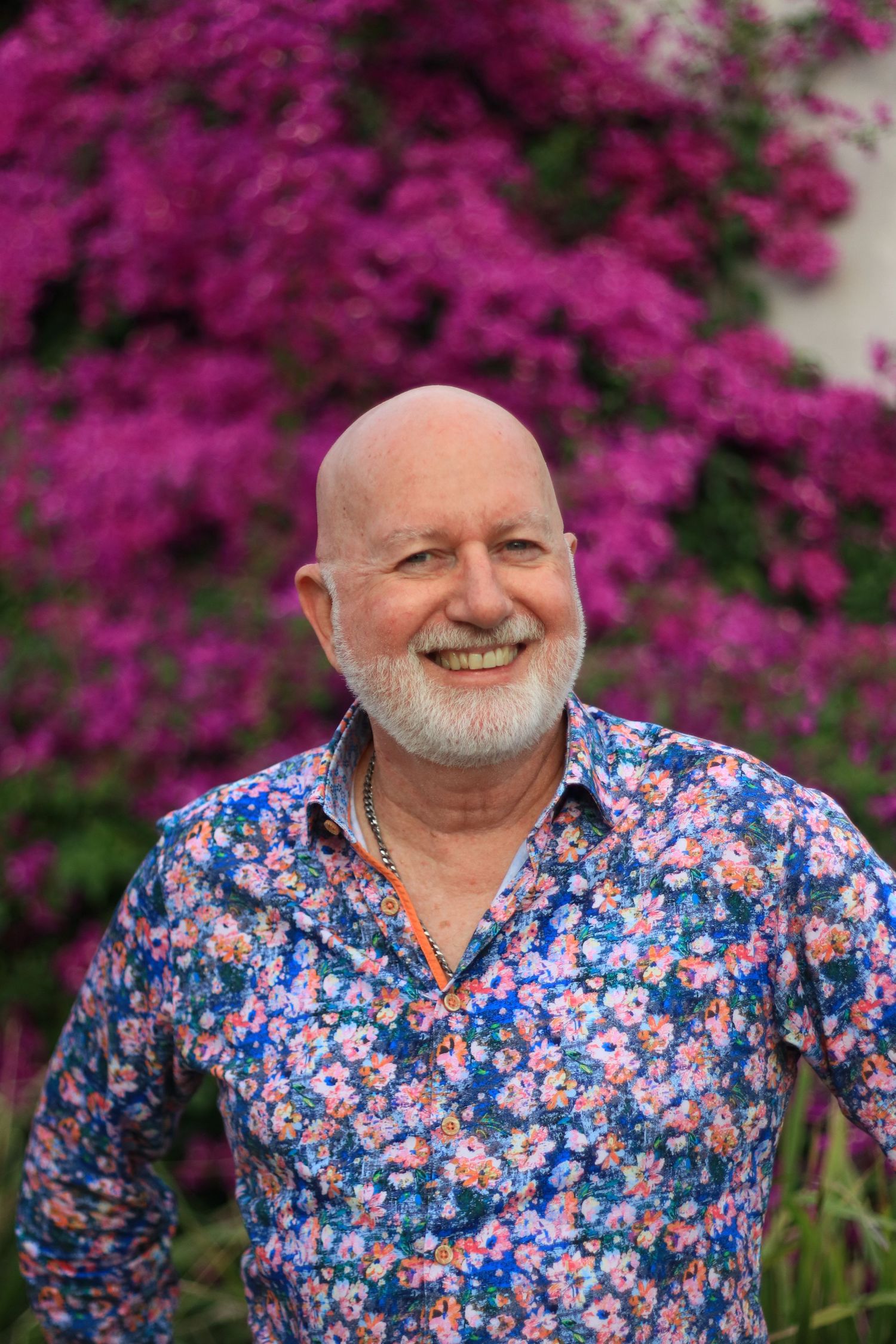 Happy bald senior man with white beard wearing floral shirt standing before purple bougainvillea.