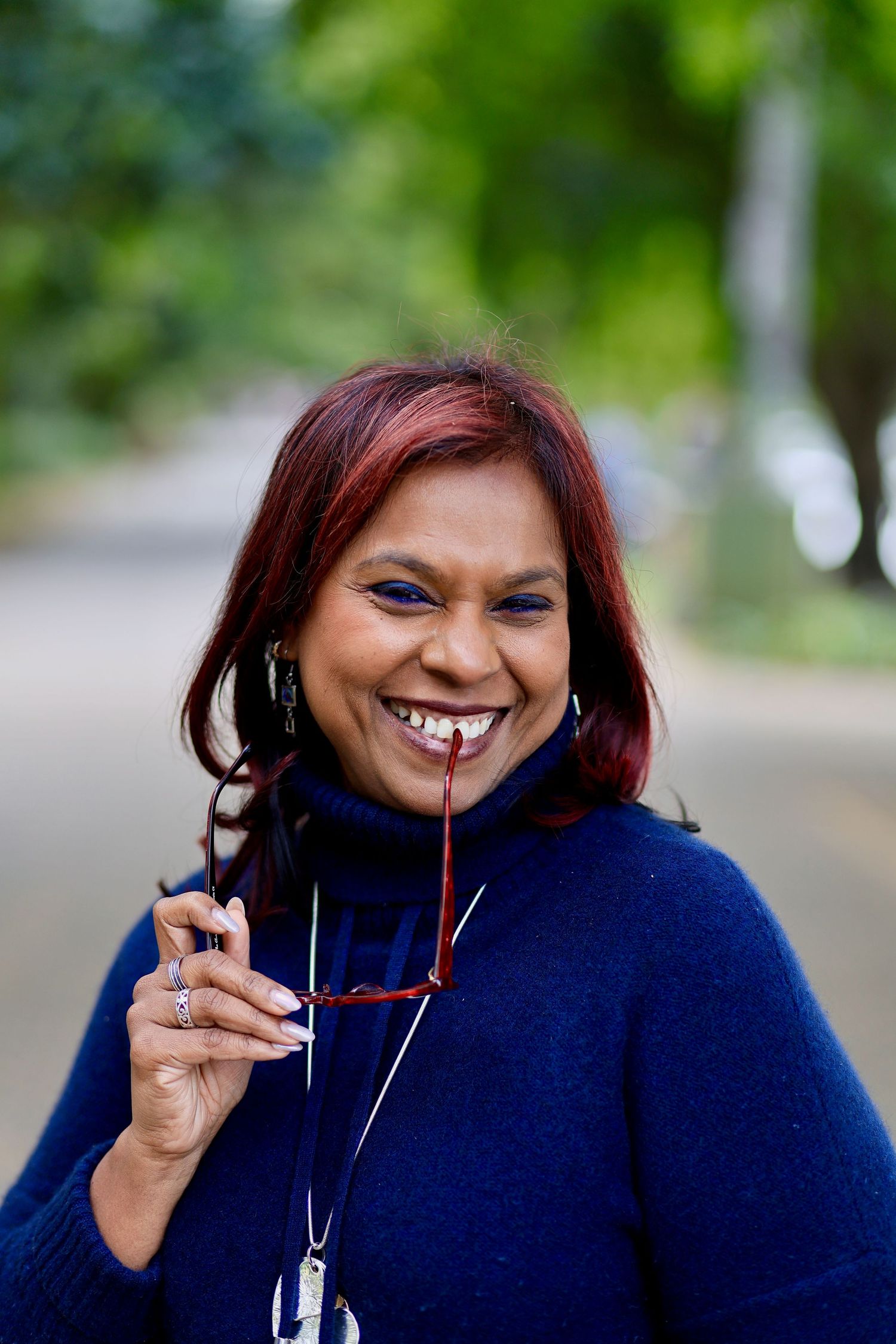 Smiling woman with red hair holding glasses outdoors, wearing a blue turtleneck sweater with trees in background.
