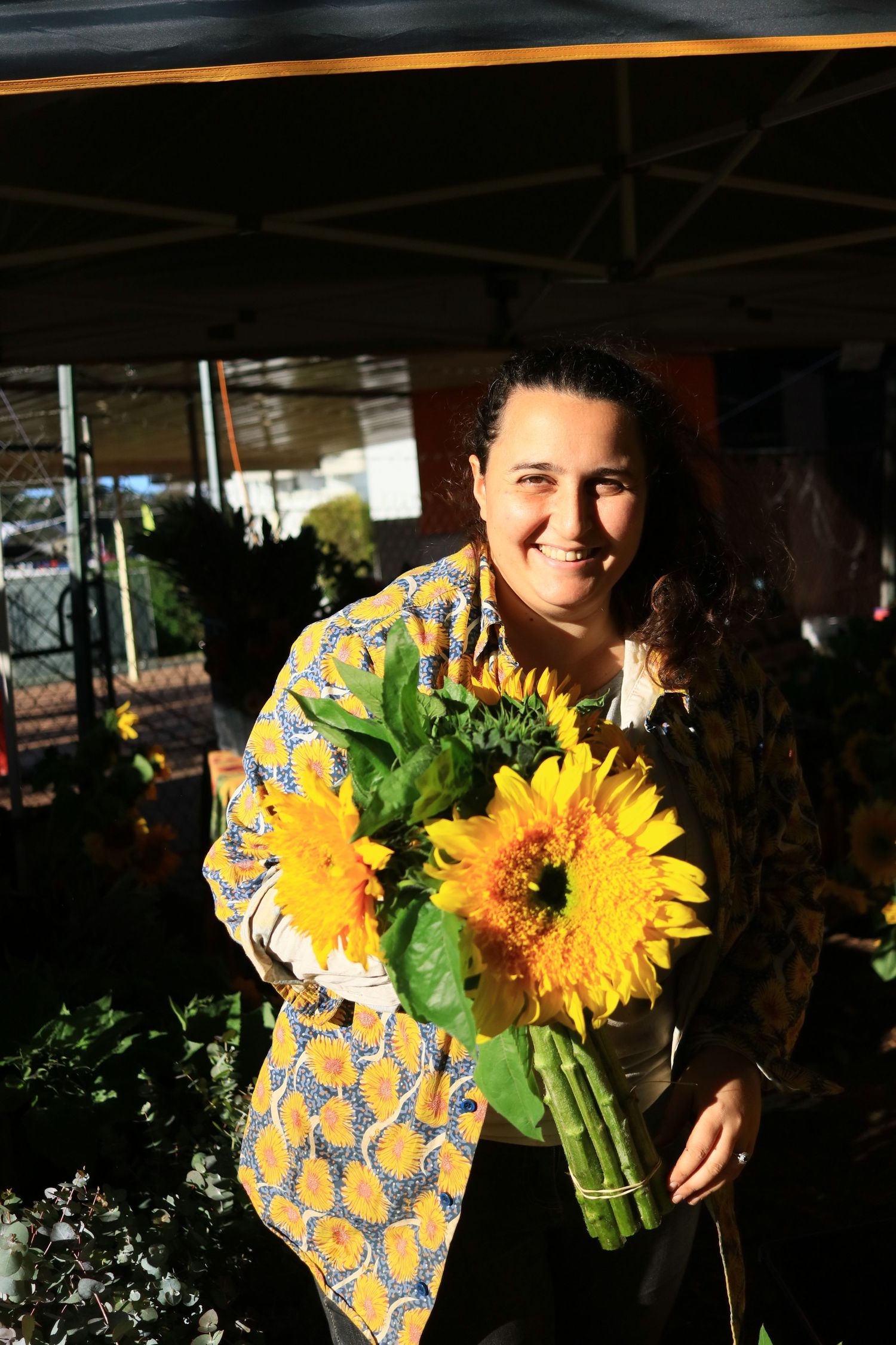 A smiling woman holding a bouquet of bright yellow sunflowers at an outdoor flower market stall.