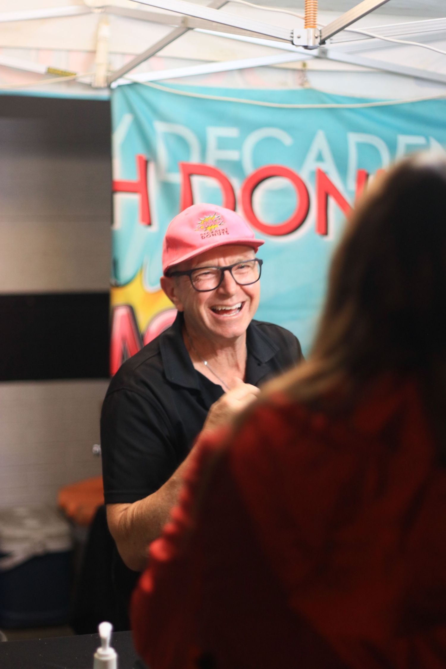 Smiling man wearing pink cap and glasses at a Decadent Donuts market stall.