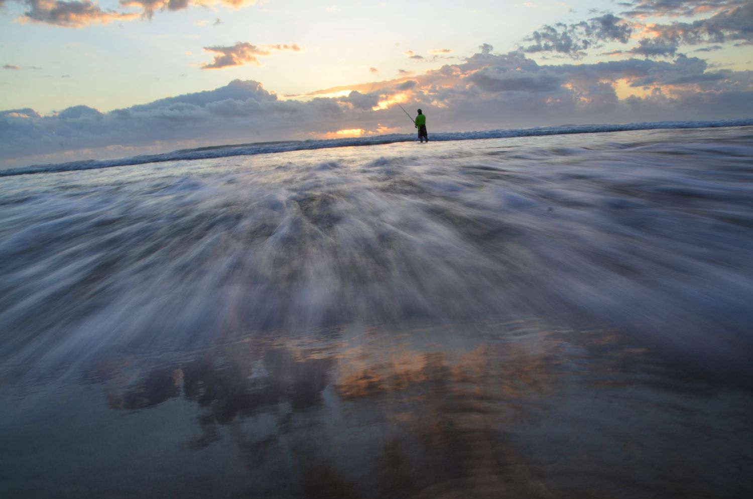 Ocean waves rushing ashore at sunset with a lone figure standing in the surf under dramatic clouds.