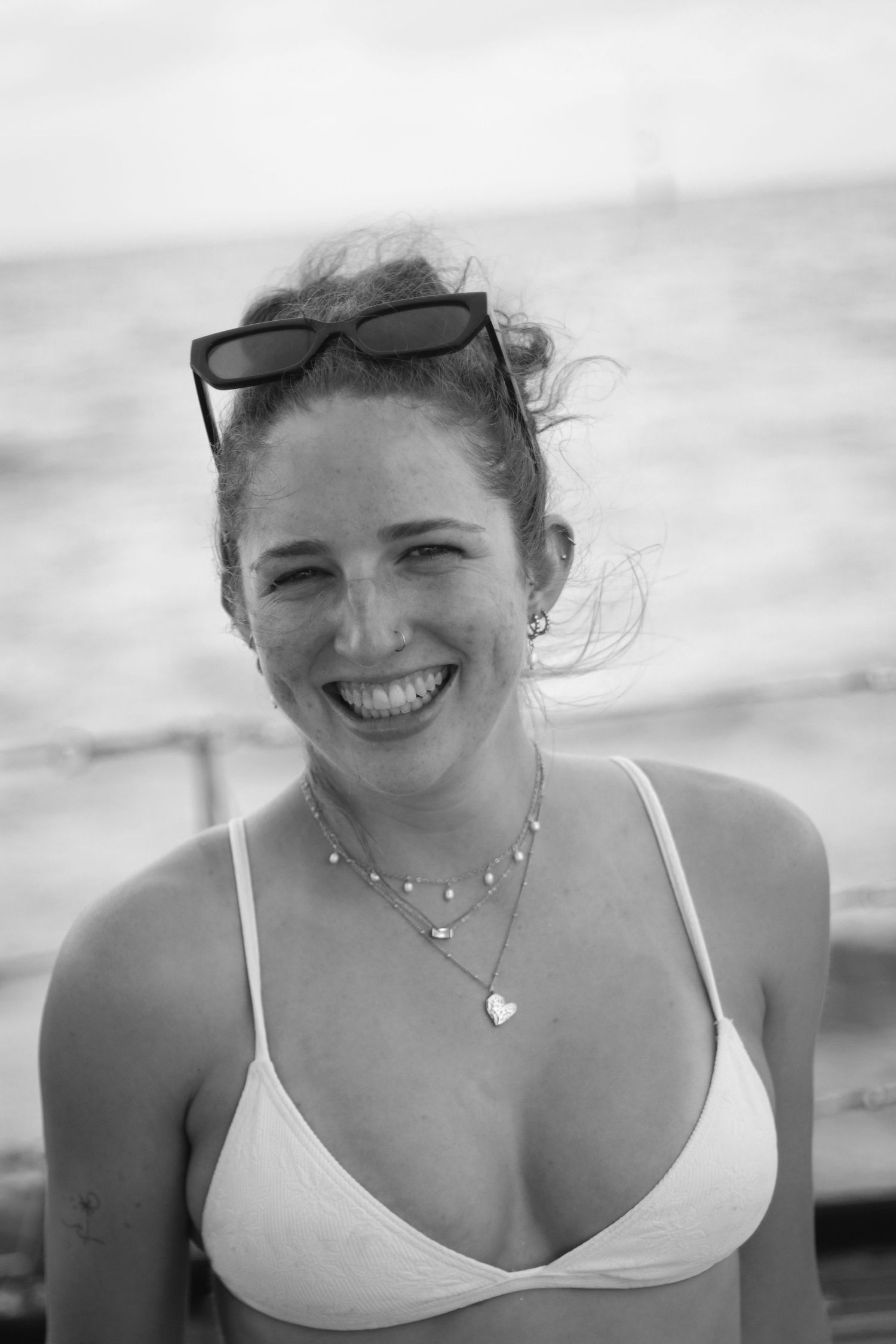 A smiling young woman in a white bikini top with sunglasses on her head, standing near the ocean in black and white.