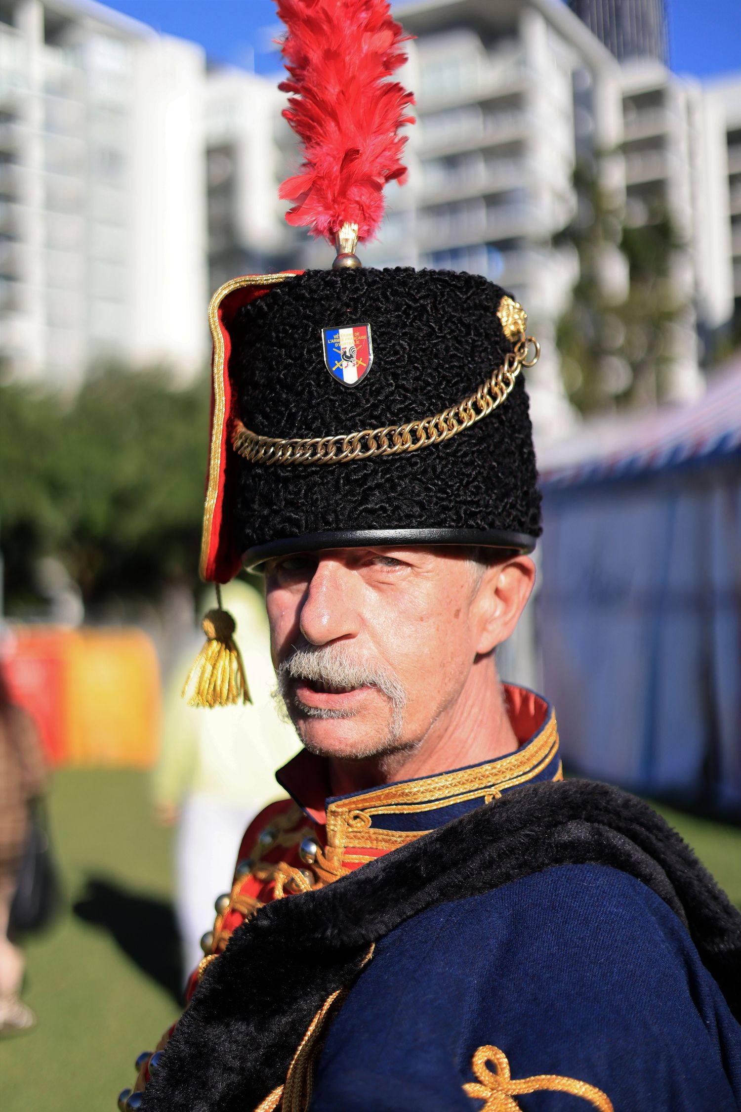 Man in French military historical costume with black fur hat, red feather plume, and blue uniform at outdoor event.