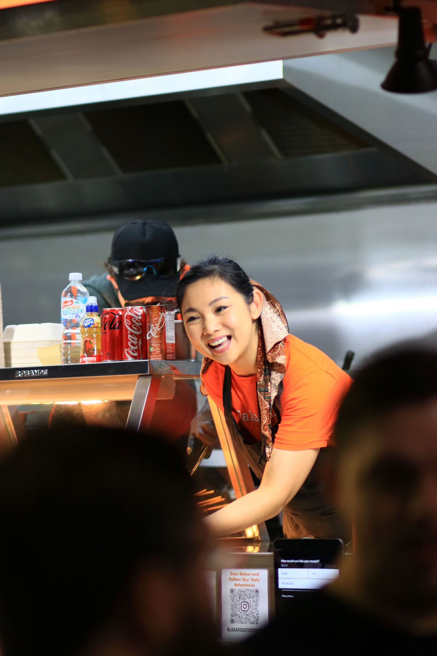 Smiling female worker in orange shirt leaning over counter in busy restaurant kitchen.