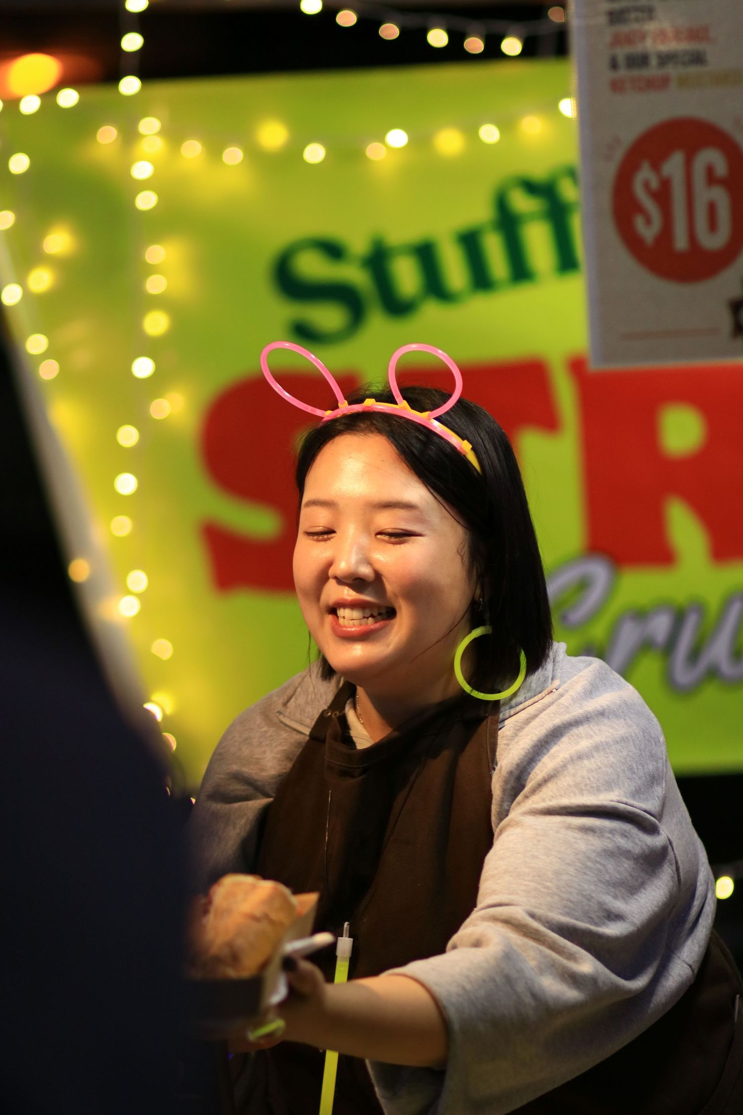 A cheerful woman wearing pink bunny ear headband and hoop earrings smiling at a Stuffed Stru food stall.