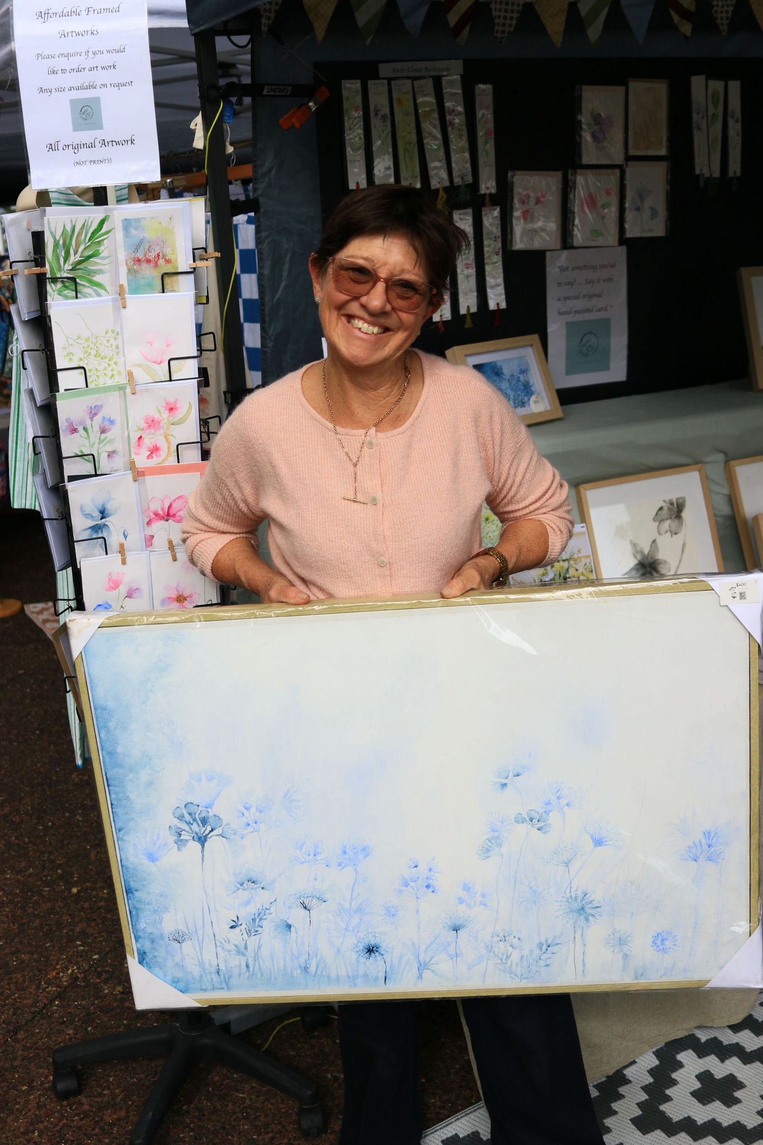A smiling woman holding a large canvas painting of blue wildflowers at an outdoor art market stall.