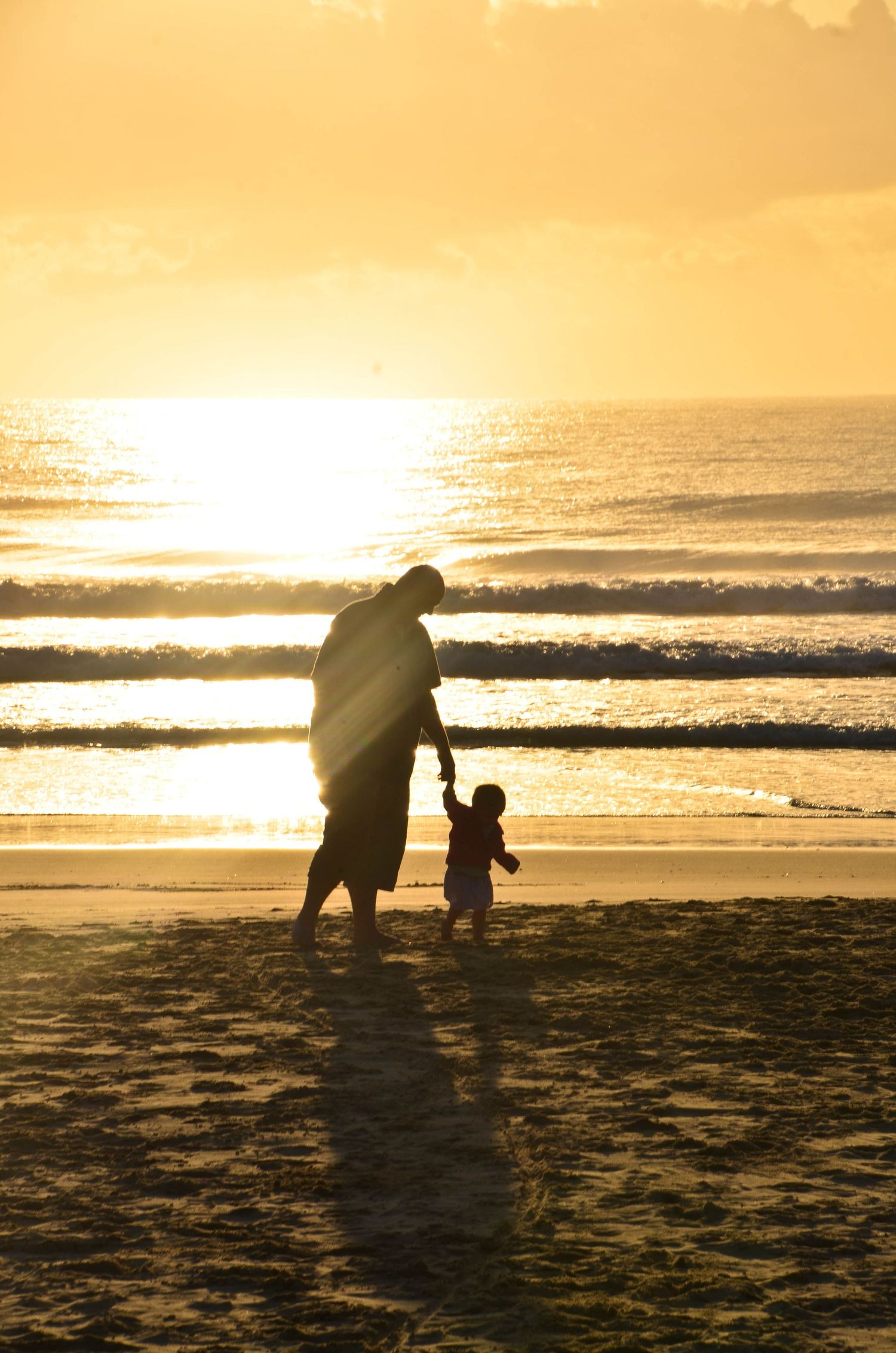A parent and toddler silhouetted against a golden sunset on a sandy beach near the ocean waves.