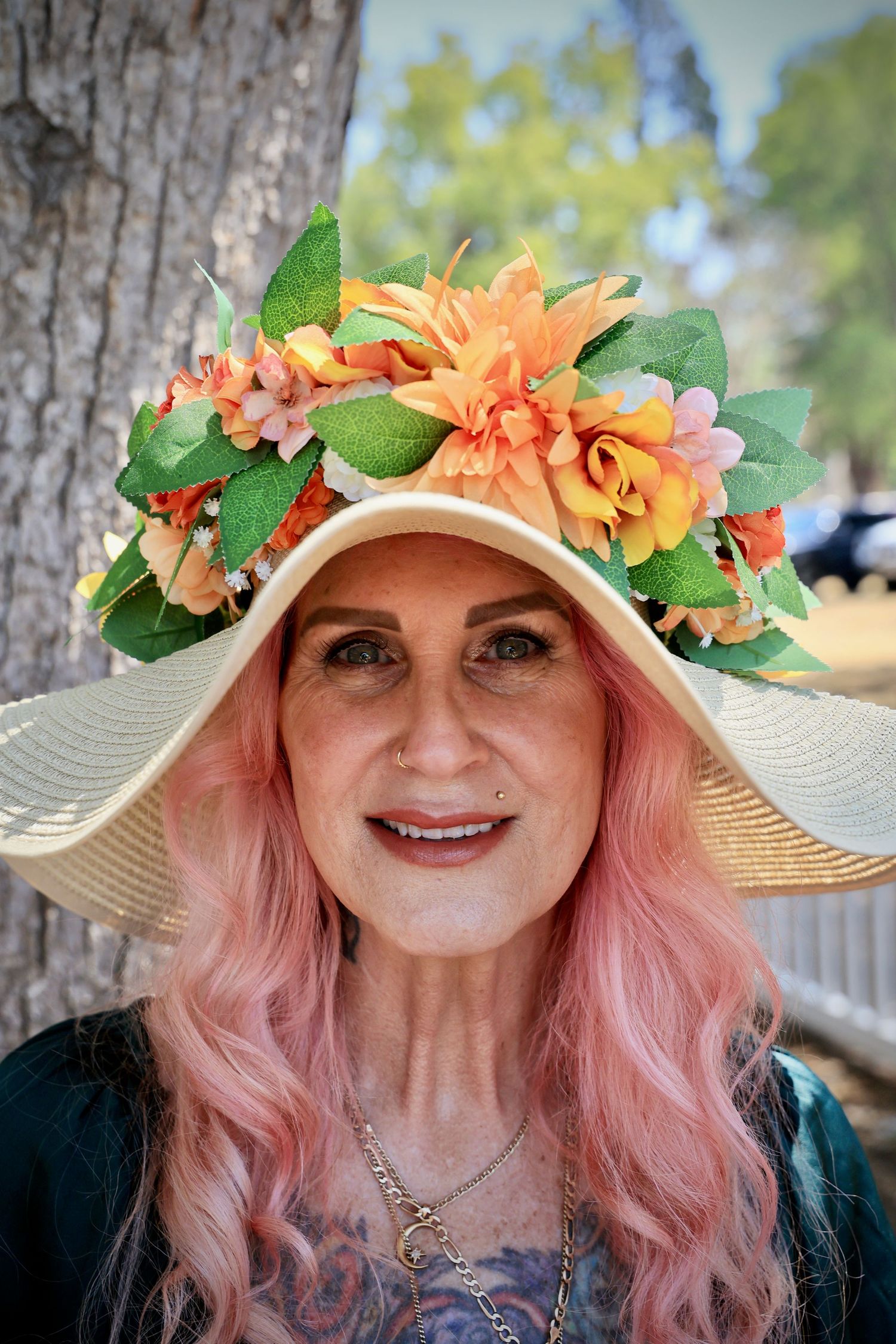 Smiling woman with pink hair wearing a wide-brim straw hat adorned with colorful orange and yellow flowers outdoors.