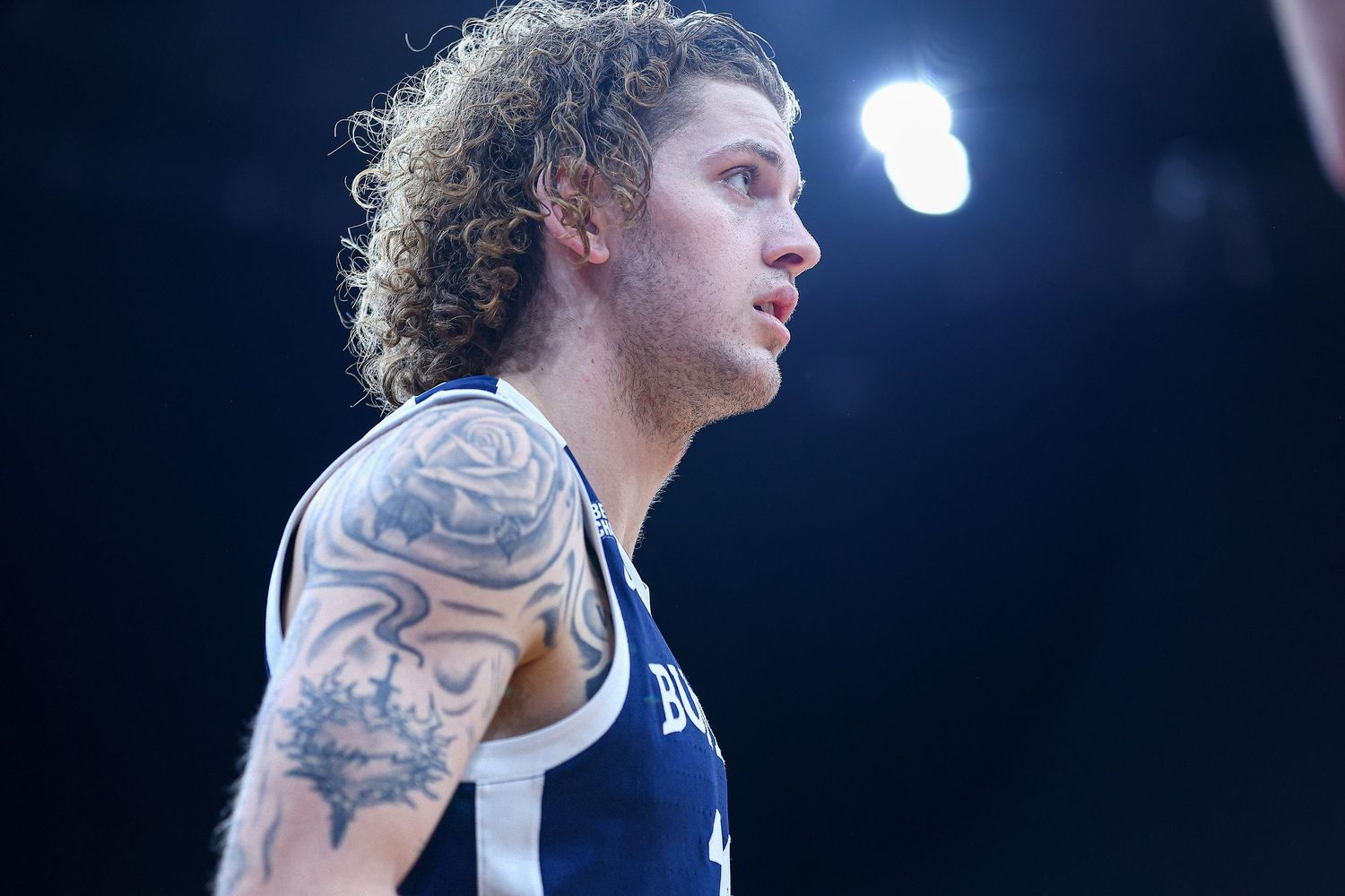 Tattooed basketball player with curly hair in blue jersey under dramatic arena lighting.