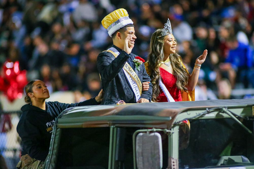 Homecoming king and queen wave from military vehicle during high school football halftime.