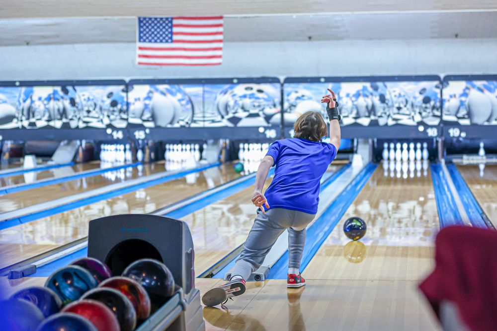 Child bowler releasing ball down a lane at an American bowling alley with US flag displayed overhead.
