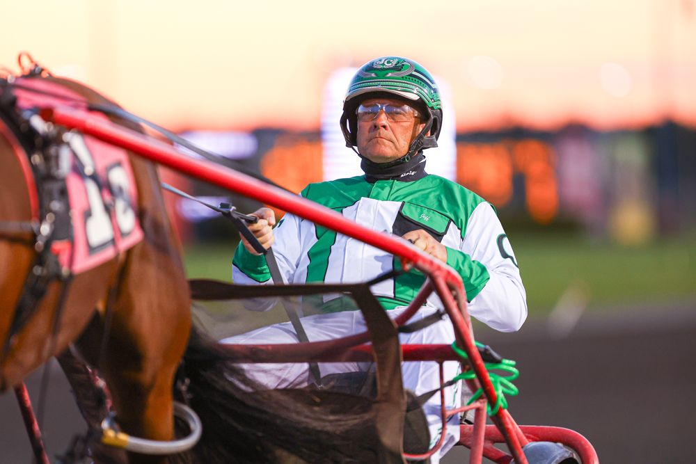 Harness racing jockey in green uniform drives sulky at dusk on dirt track.