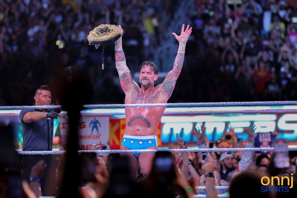 Tattooed WWE champion raises arms triumphantly holding title belt before a massive cheering arena crowd.