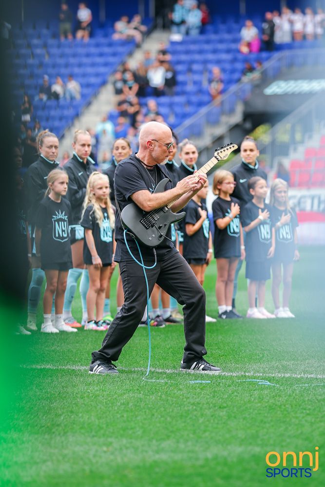 Bald guitarist performs electric guitar on stadium field during pre-game ceremony with children lined up.