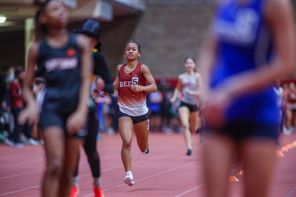Female track athlete in red Bees uniform running indoor race on red track.