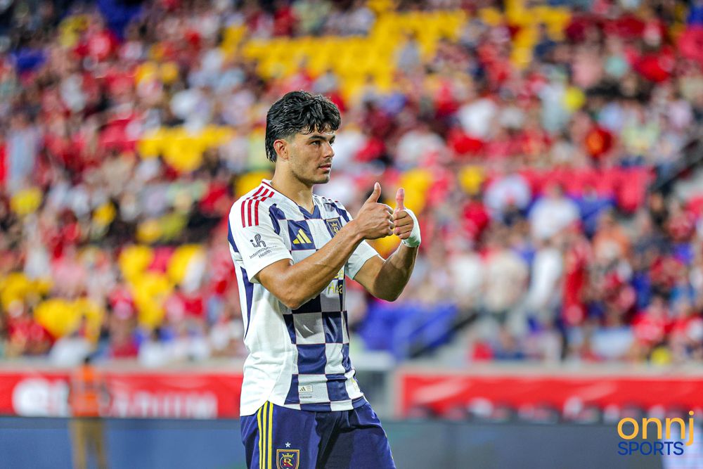 Soccer player in blue and white checkered jersey applauds fans during an MLS match.