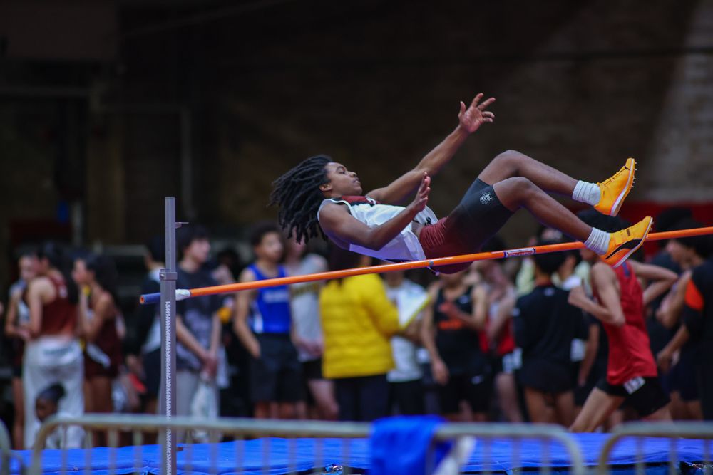 Athlete performs high jump clearing bar at indoor track and field competition with crowd behind.