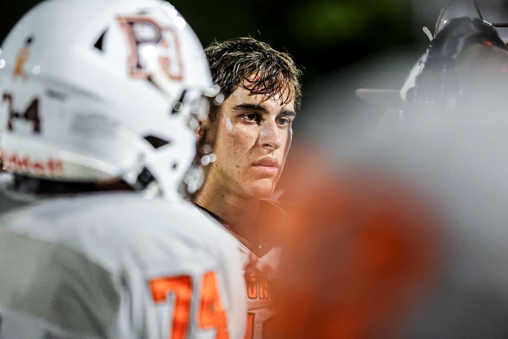 High school football player in orange PJ uniform looking focused on sideline during night game.