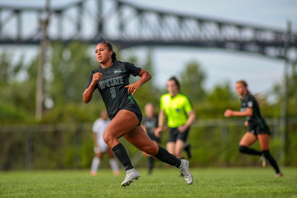 Upstate University Female soccer player in black uniform sprints across green field during competitive match with bridge in background.