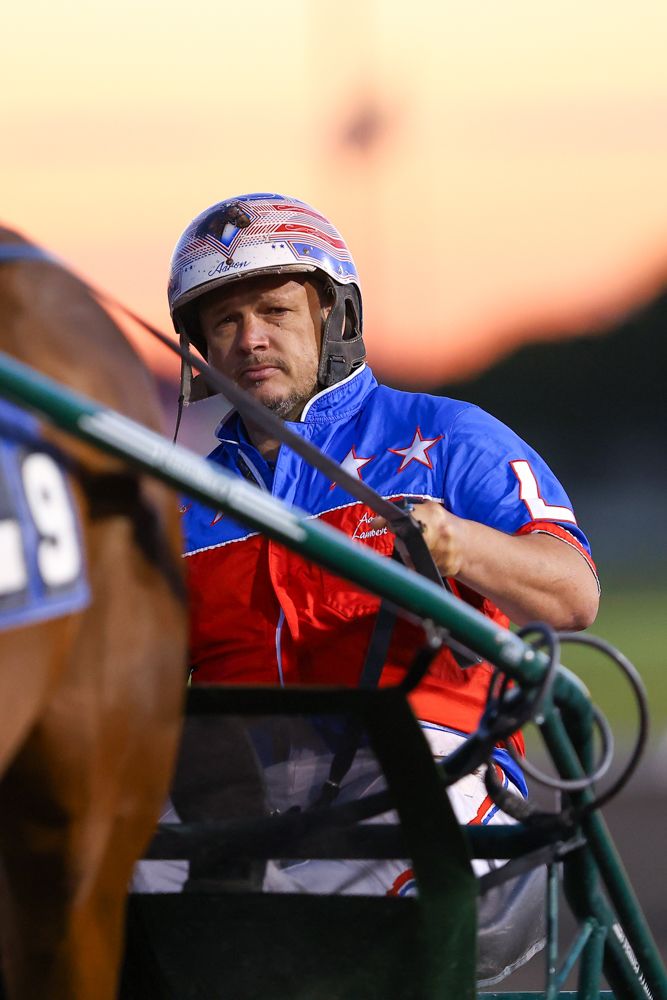 A harness racing jockey in a blue and red star-spangled jersey and helmet guides his horse at sunset.