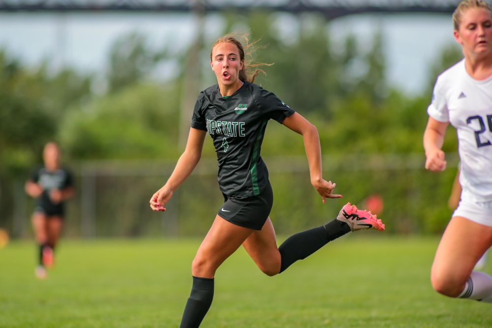 Female soccer player in green uniform kicking ball during competitive college soccer match outdoors.