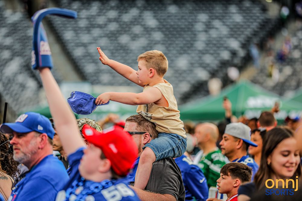 Young child sits on an adults shoulders waving a blue cap at a New York Giants NFL fan event.