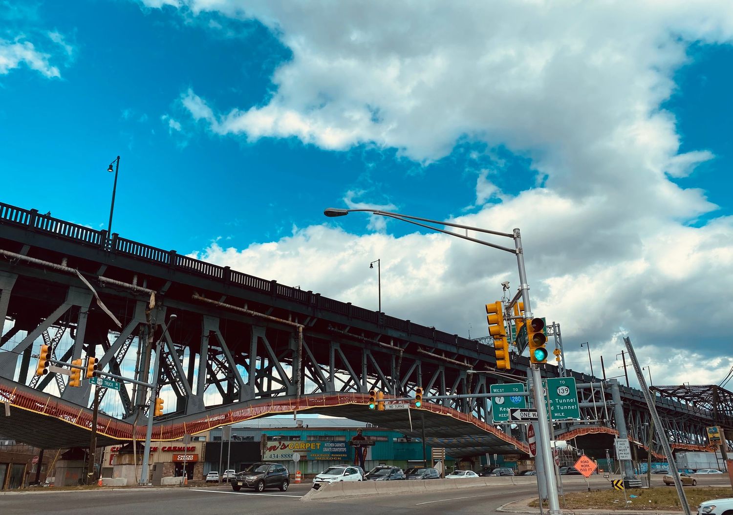 A steel bridge overpass with traffic signals and green highway signs under a partly cloudy blue sky.