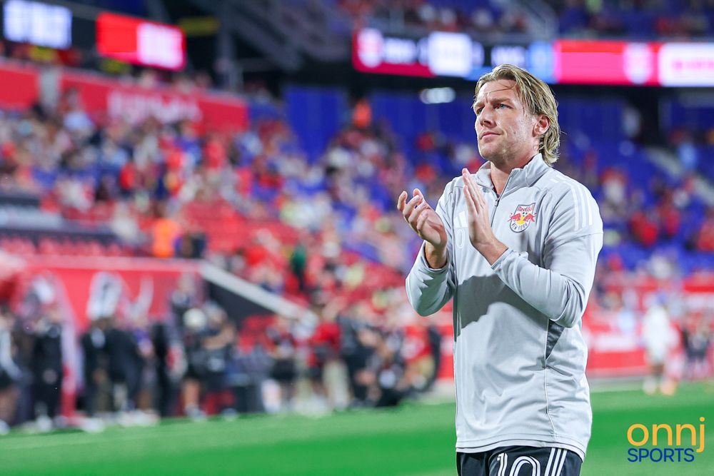 New York Red Bulls player in grey jacket applauds fans in a packed soccer stadium.