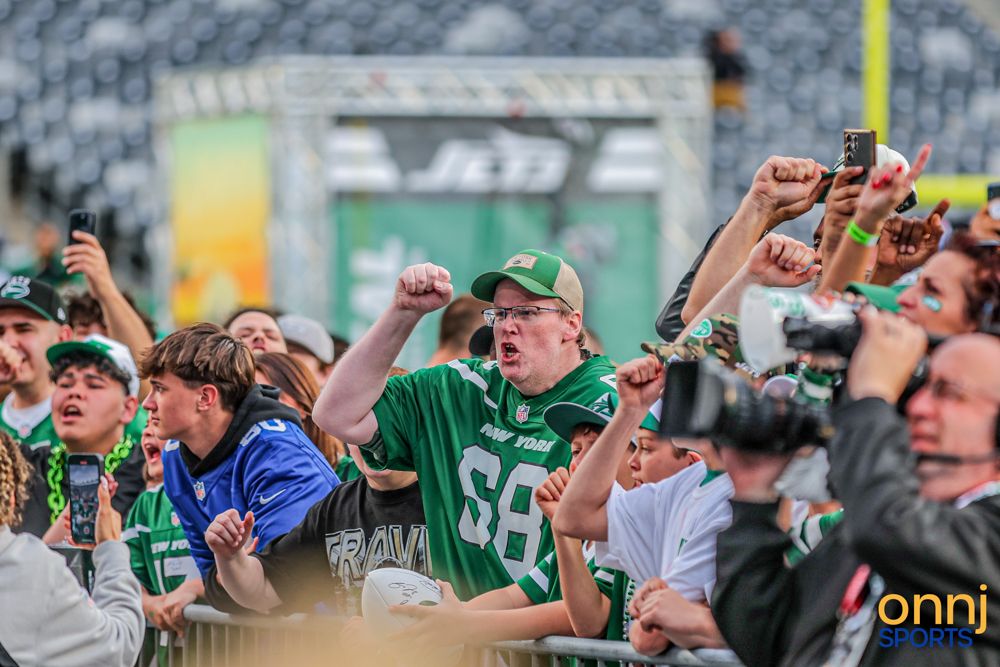 Enthusiastic New York Jets fans in green jerseys cheer behind a barrier at a stadium event.