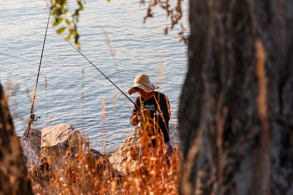 A fisherman in a straw hat stands on rocky shore casting a fishing line into calm blue water at golden hour.