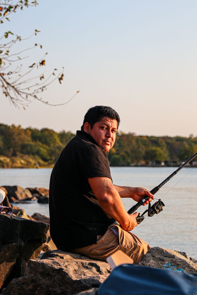 A man in a black shirt sits on rocks holding a fishing rod by a calm river at sunset.