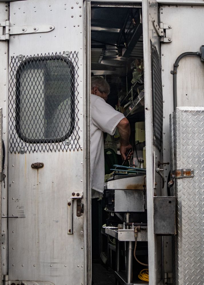A cook in a white uniform works inside a narrow stainless steel food truck kitchen.
