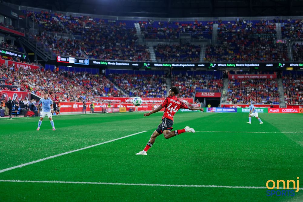 Red-uniformed soccer player dribbles ball toward goal on a bright green stadium pitch.