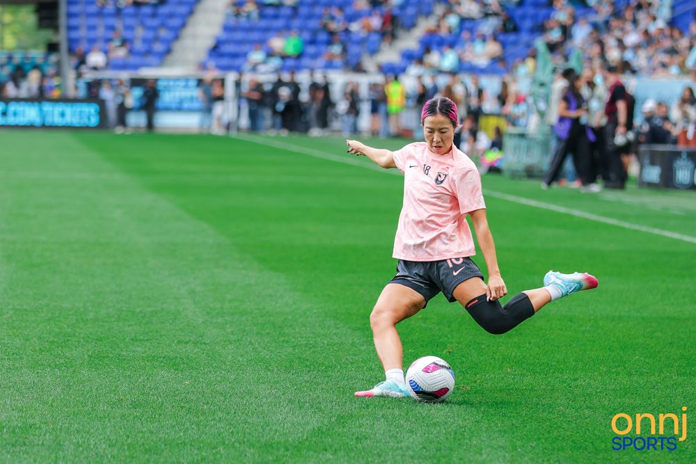 Female soccer player in pink jersey practices kicking during warmups on a professional soccer field.