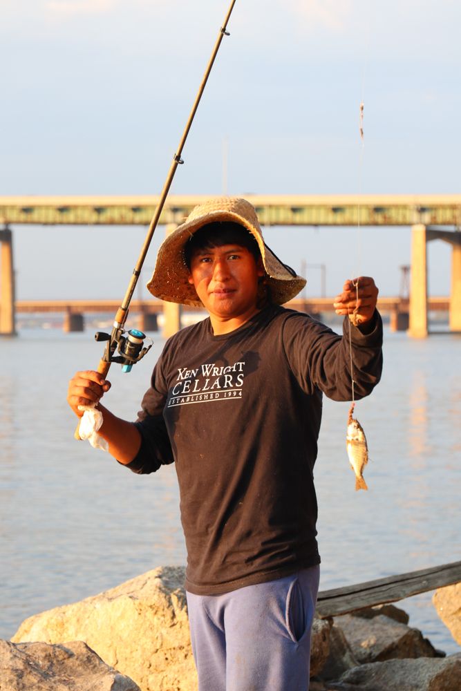 Fisherman in straw hat proudly displaying freshly caught fish by a waterway bridge at sunset.