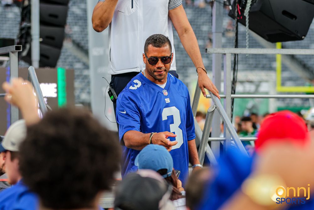 Man wearing a New York Giants number 3 jersey signs autographs for fans at a stadium event.