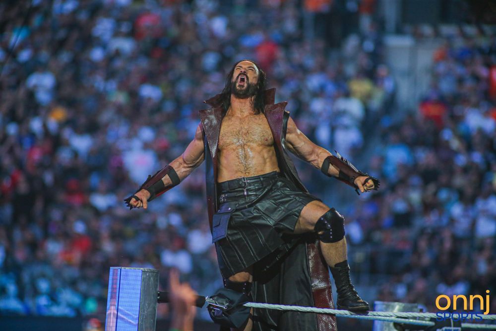 WWE wrestler Drew McIntyre poses triumphantly on ring ropes before a massive crowd at event.
