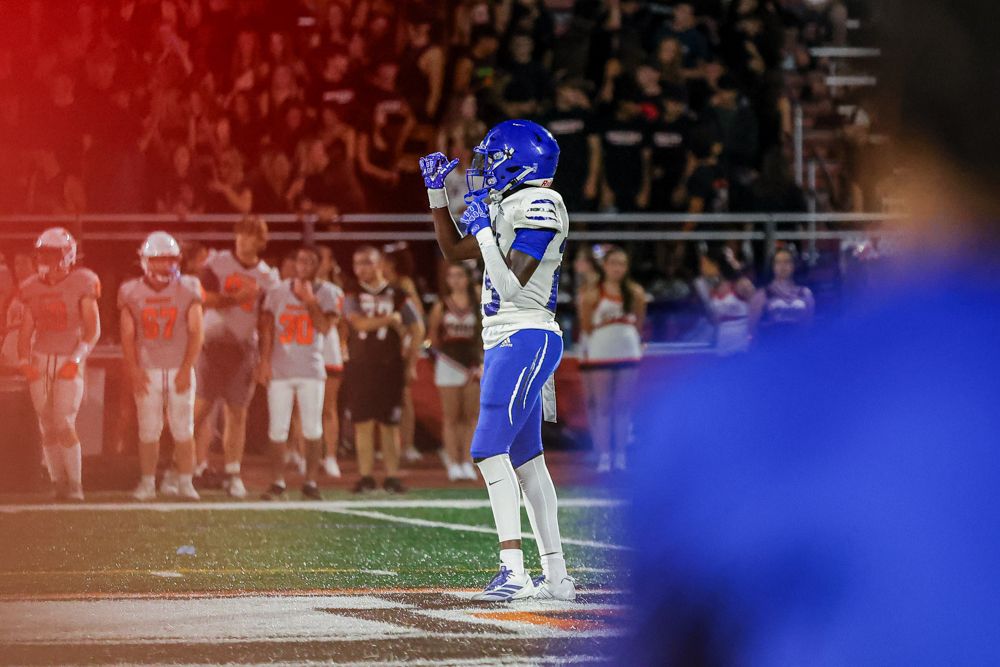 Football player in blue uniform raises helmet overhead in celebration during night game on field.
