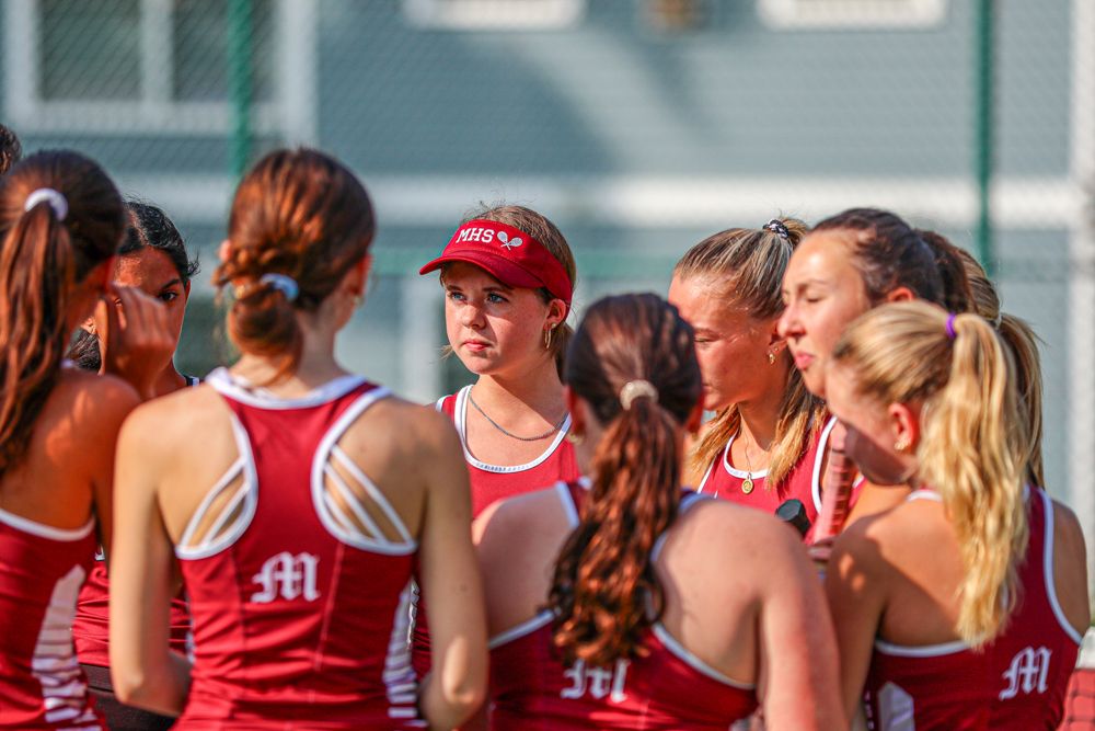 Girls tennis team in red uniforms huddling together with coach before outdoor match.