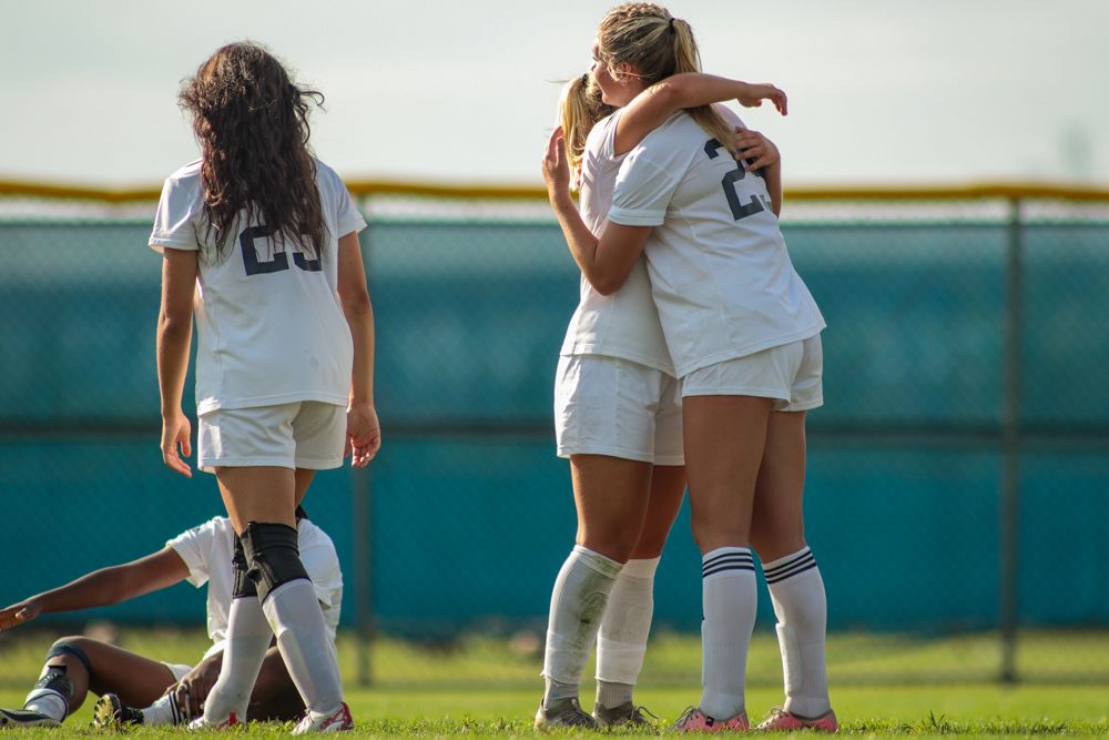Two female soccer players embrace in celebration during outdoor high school soccer game.