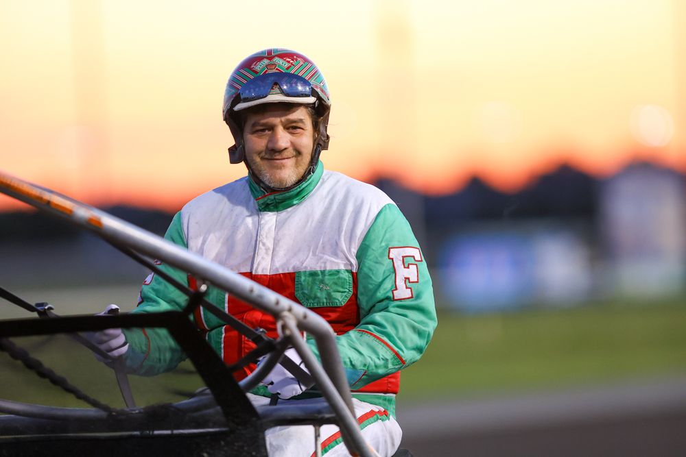 Smiling harness racing jockey in green and white silks poses in sulky at sunset on a racetrack.