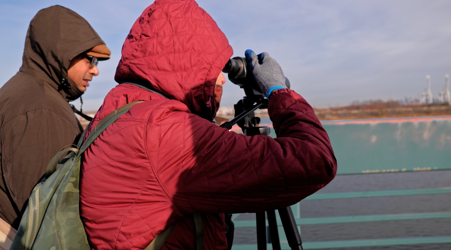 Two people in winter jackets use a camera on a tripod to photograph scenery from a bridge.