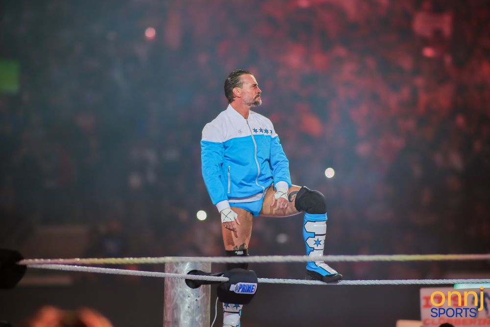WWE wrestler perches on ring ropes in blue jacket during dramatic arena entrance.