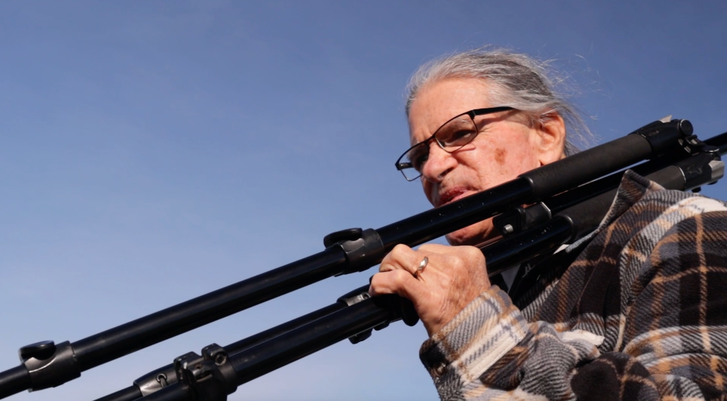 Close-up of senior photographer looking through camera lens mounted on tripod against blue sky.