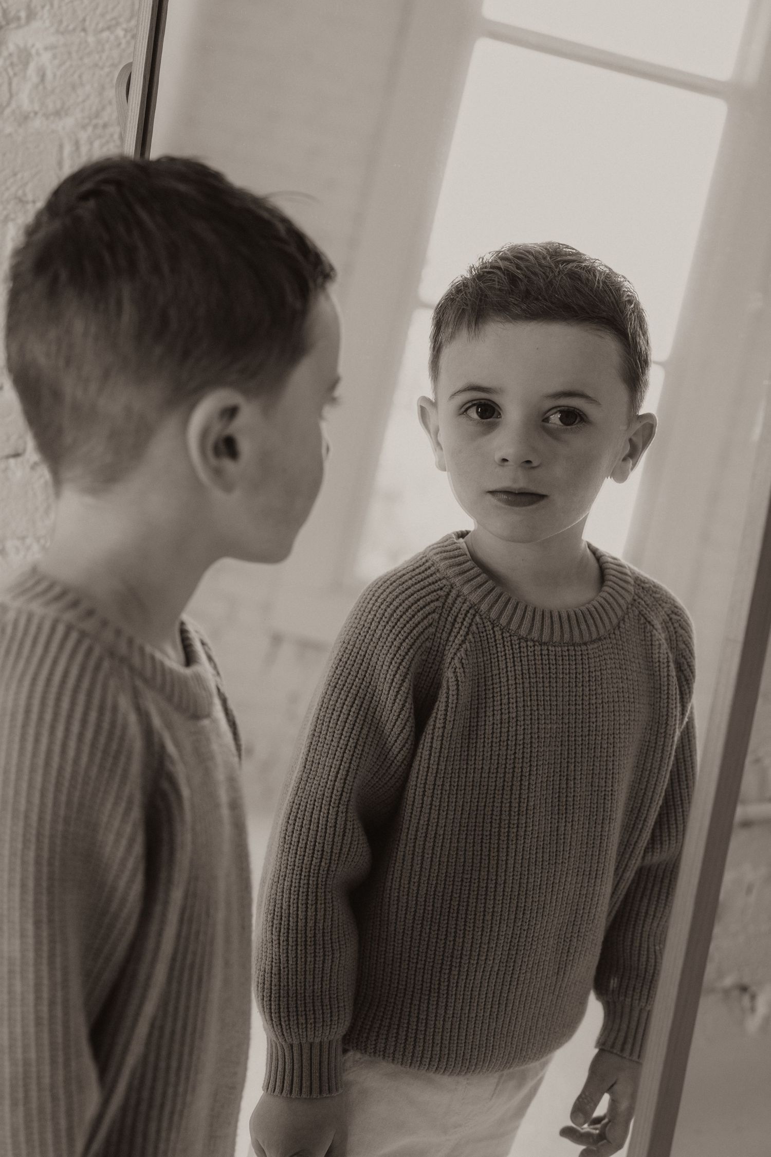 A young boy in a knit sweater gazes at his reflection in a mirror, captured in sepia tone.